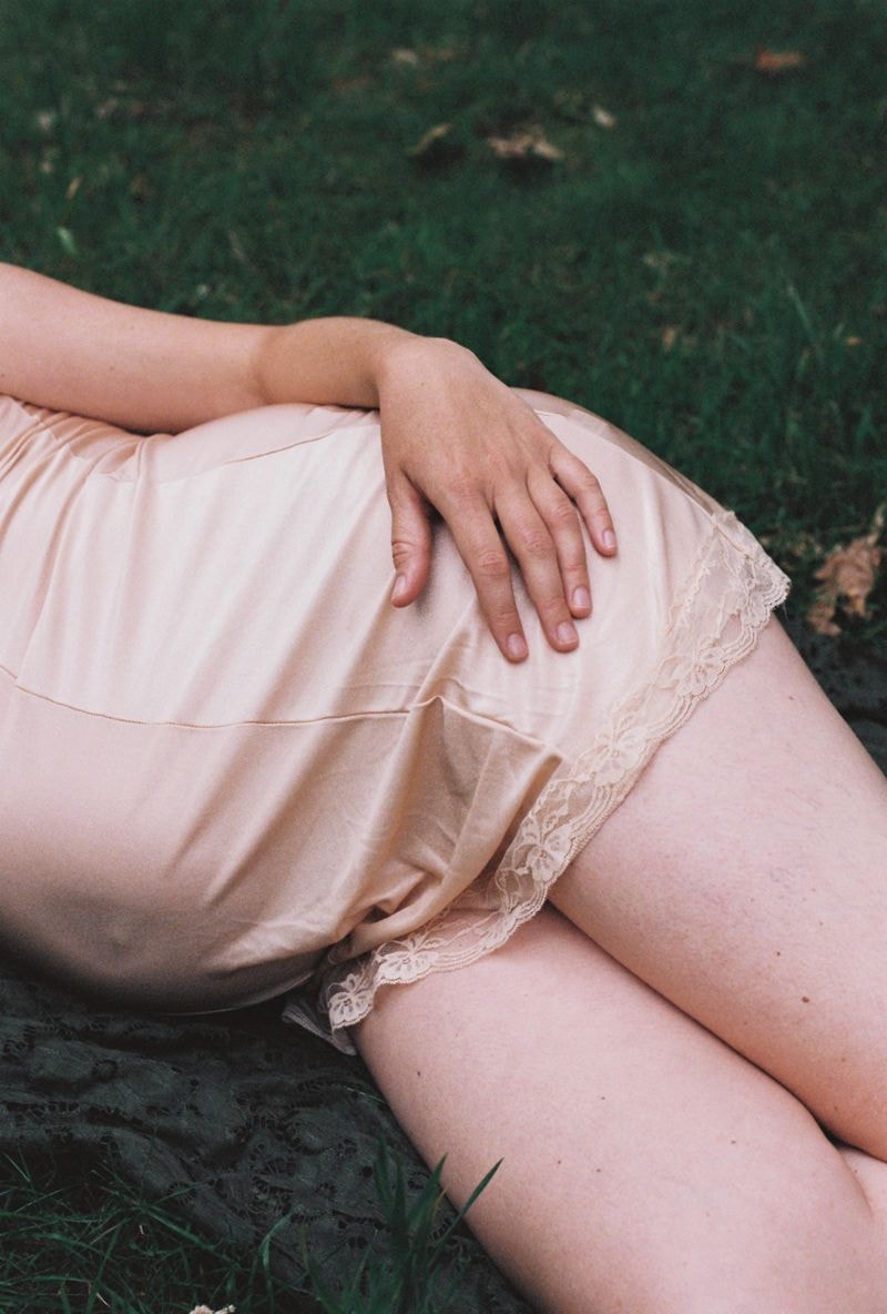 Zwangere vrouw met hand op heup staat in grasveld, focus op ronde buik en natuurlijke omgeving